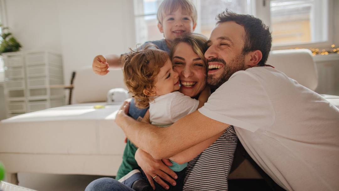 Protect Your Family with Life Insurance. Family hugging in front of the sofa in their living room.